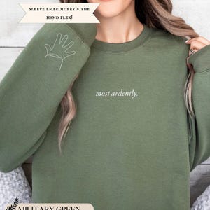 Puede incluir: Sudadera verde militar con la frase "most ardently" en escritura blanca. La manga izquierda presenta un contorno de mano blanco. El texto "SLEEVE EMBROIDERY - THE HAND FLEX!" est&aacute; encima de la sudadera. La parte inferior de la imagen tiene el texto "MILITARY GREEN".