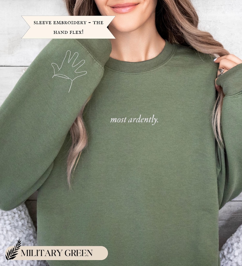 Puede incluir: Sudadera verde militar con la frase "most ardently" en escritura blanca. La manga izquierda presenta un contorno de mano blanco. El texto "SLEEVE EMBROIDERY - THE HAND FLEX!" est&aacute; encima de la sudadera. La parte inferior de la imagen tiene el texto "MILITARY GREEN".