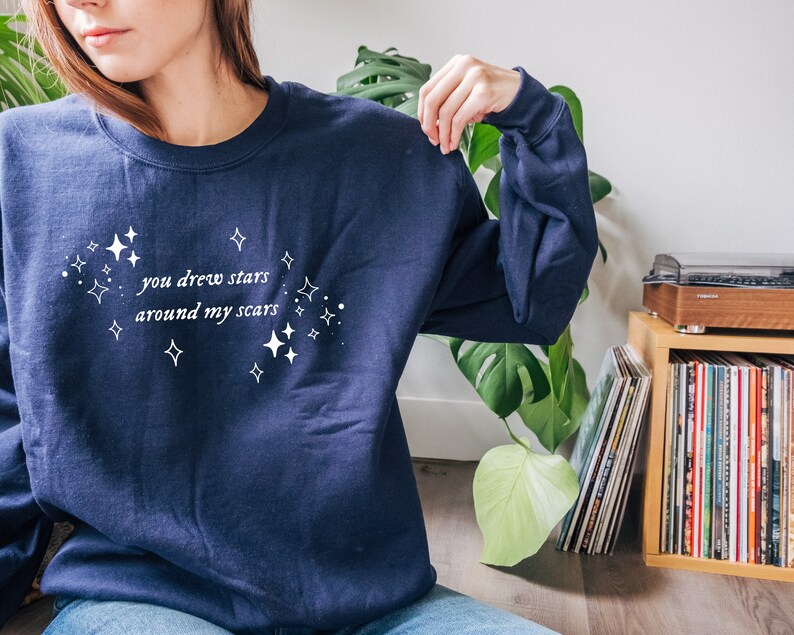 May include: A navy blue sweatshirt with the text "you drew stars around my scars" in white lettering and a white star pattern.