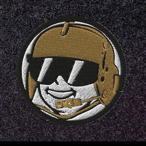 May include: Embroidered patch featuring a smiling pilot wearing a gold helmet and sunglasses. The patch has a black border and is stitched with gold, white, and black thread. The pilot's face is detailed with white and black stitching.