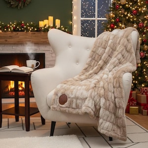 May include: A beige faux fur throw blanket draped over a white armchair. The blanket has a textured, quilted pattern and a brown leather patch. The scene includes a Christmas tree, a fireplace, and a small side table with a cup of tea.