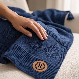 May include: A navy blue knitted throw blanket featuring a textured diamond pattern and a tassel at the corner. A circular tan leather patch is attached to the corner, displaying the letter R enclosed within a decorative laurel wreath design.
