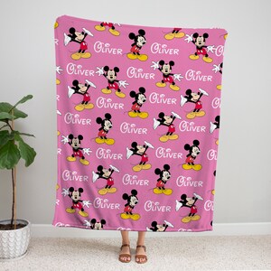 May include: Pink fleece blanket with a repeating pattern of Mickey Mouse in a red and yellow outfit waving. The name "Oliver" is repeated throughout the pattern.