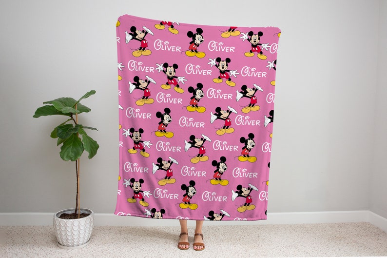 May include: Pink fleece blanket with a repeating pattern of Mickey Mouse in a red and yellow outfit waving. The name "Oliver" is repeated throughout the pattern.
