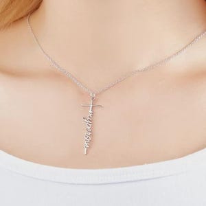 Könnte beinhalten: Eine silberfarbene Halskette mit einem Kreuzanhänger. Der Anhänger trägt das Wort "inspired" vertikal in Schreibschrift. Die Halskette wird um den Hals getragen.