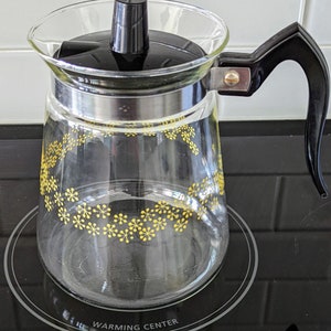 May include: A glass coffee pot with a black lid and handle. The pot has a yellow and white floral pattern and the words "Warming Center" printed on the bottom.