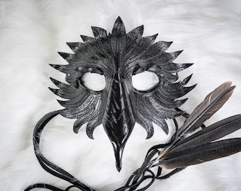 Puede incluir: Una máscara de cuero negro con un diseño de pájaro, con un pico largo y detalles de plumas. La máscara tiene aberturas para los ojos y está adornada con plumas negras adjuntas y lazos de cuero para asegurar. La máscara está sobre un fondo blanco y esponjoso.
