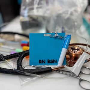 Puede incluir: Una placa azul turquesa RN BSN con una cadena plateada y un rotulador azul y blanco. En el fondo hay equipo médico, como un estetoscopio y unas tijeras. La placa es rectangular y tiene el texto "RN BSN" impreso.