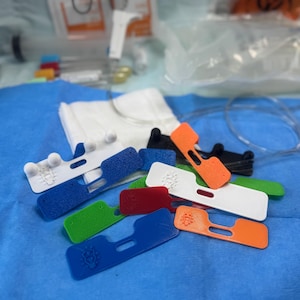 May include: A collection of colorful, 3D-printed medical device components. The components are in various colors, including white, blue, green, red, black, and orange. Each component has a unique shape with a small logo.