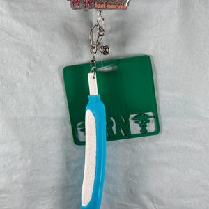May include: A badge reel with a glittery brain design that says "Oh look! It's my last nerve." The reel has a blue and white pen-like object attached, along with a green RN badge holder.