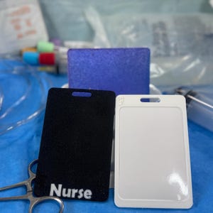 May include: A black badge holder with the word "Nurse" in white text, a white badge holder, and a blue glittery rectangle. The badge holders have a slot at the top for a lanyard. Medical supplies are in the background.