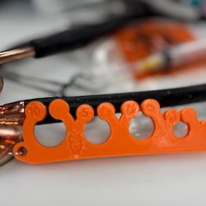 May include: An orange 3D-printed tool with multiple circular openings and numbered markings, likely for medical use. The tool is positioned near a stethoscope and other medical supplies, suggesting a healthcare context.