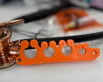 May include: An orange 3D-printed tool with multiple circular openings and numbered markings, likely for medical use. The tool is positioned near a stethoscope and other medical supplies, suggesting a healthcare context.