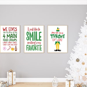 May include: Three framed prints with festive text in red and green, perfect for Christmas. The prints are displayed against a grey wall, with a white Christmas tree and gifts in the background. The text includes phrases like "We Elves" and "Smile".