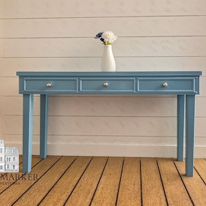 May include: A miniature blue console table featuring three drawers with small silver knobs. A white vase containing a small white and blue flower arrangement sits on the center of the table surface. The table has four straight legs.