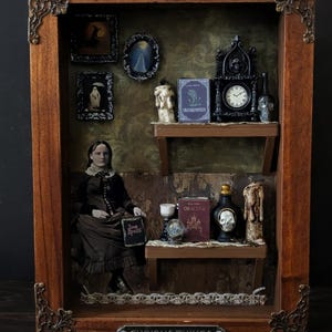 May include: A wooden shadow box with ornate metal corner accents and a plaque reading CURIOUS THINGS. Inside are two shelves holding a miniature Frankenstein book, a skull clock, a Dracula book, a crystal ball, and a figure holding a Love Spells sign.