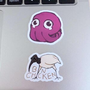 K&ouml;nnte beinhalten: Zwei Aufkleber auf einem Laptop. Der obere Aufkleber ist ein lila Cartoon-Oktopus mit gro&szlig;en Augen. Der untere Aufkleber ist ein Cartoon-Vogel mit den Worten "BIN CHICKEN" darauf.