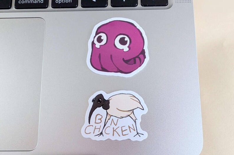 K&ouml;nnte beinhalten: Zwei Aufkleber auf einem Laptop. Der obere Aufkleber ist ein lila Cartoon-Oktopus mit gro&szlig;en Augen. Der untere Aufkleber ist ein Cartoon-Vogel mit den Worten "BIN CHICKEN" darauf.