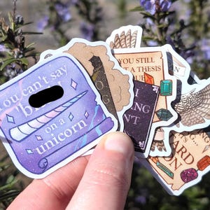 May include: A collection of die-cut vinyl stickers featuring various designs, including a purple scroll with the text 'You can't say [redacted] on a unicorn', a dark book, a scroll with the text 'You still got a thesis due', and a pair of wings.