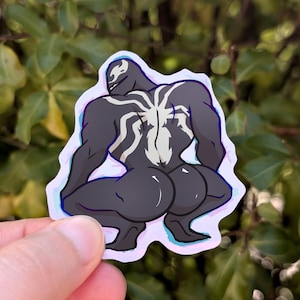 Könnte beinhalten: Ein Aufkleber mit einer Cartoon-Illustration eines muskulösen, dunkelgrauen Charakters in einer hockenden Pose. Die Figur hat ein weißes spinnenartiges Design auf dem Rücken und eine weiße Umrandung. Der Aufkleber wird vor einem Hintergrund aus grünen Blättern gehalten.