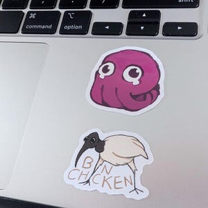 K&ouml;nnte beinhalten: Zwei Aufkleber auf einem Laptop. Einer ist ein Cartoon-Oktopus in Rosa mit gro&szlig;en Augen. Der andere ist ein Cartoon-Vogel mit dem Text "BIN CHICKEN".