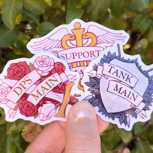 May include: Three colorful stickers with a gaming theme. One features a crown and wings with the word "SUPPORT". Another has roses and the words "DPS MAIN". The third has a shield and the words "TANK MAIN".