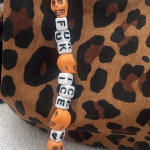 Puede incluir: Un llavero de cuentas con cuentas de calavera naranja y cuentas de letras blancas que deletrean "FUCK ICE". El llavero está colocado sobre un fondo con estampado de leopardo.