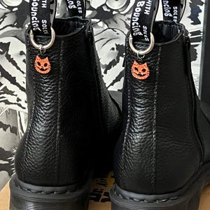 Puede incluir: Un par de botines de cuero negro con cremallera lateral. Las botas tienen una superficie texturizada y una suela de goma negra. Las botas tienen un pequeño amuleto de gato naranja sujeto a los cordones. Los cordones tienen una etiqueta que dice "With Bouncing Soles".