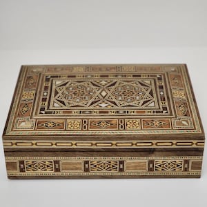 Puede incluir: Una caja de madera con intrincados patrones de incrustaciones en varios tonos de marrón, beige y negro. La caja tiene una tapa con bisagras y un diseño decorativo en la parte superior e inferior.