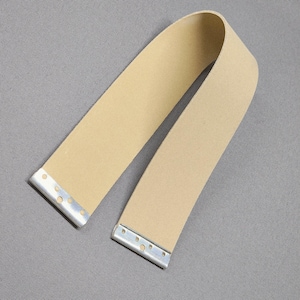May include: A tan-colored strap with metal end pieces. The strap is bent in a curved shape, and the metal ends have small holes. The background is a solid gray color.