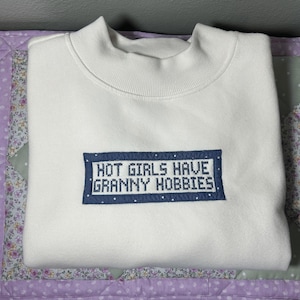 Puede incluir: Una sudadera blanca de cuello redondo con un parche rectangular bordado en el centro. El parche es azul con lunares blancos y contiene el texto HOT GIRLS HAVE GRANNY HOBBIES bordado en una fuente blanca estilo punto de cruz.