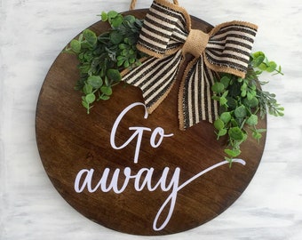 May include: A round wooden door hanger with a black and white striped bow and green foliage. The door hanger has the words "Go away" written in white cursive lettering.