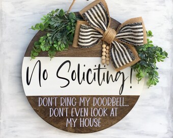 May include: A round wooden door hanger with a white and brown striped bow, green foliage, and the text "No Soliciting! Don't ring my doorbell... Don't even look at my house".