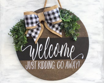 May include: A round wooden door sign featuring a black and brown horizontal split design. The text reads "Welcome" in white script and "JUST KIDDING, GO AWAY!" in white block letters. It is decorated with a buffalo plaid bow and greenery.