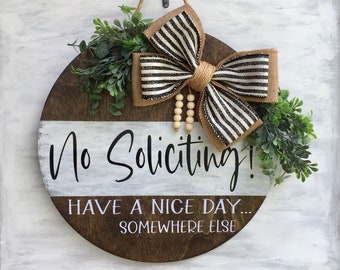 May include: A round wooden sign with the text "No Soliciting! Have a Nice Day... Somewhere Else" in white lettering. The sign is decorated with a black and white striped burlap bow and green foliage.