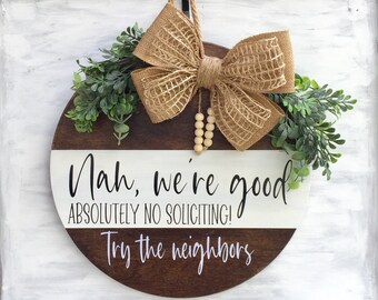 May include: A round wooden sign with the text "Nah, we're good. Absolutely no soliciting! Try the neighbors." The sign is decorated with a burlap bow and green foliage.