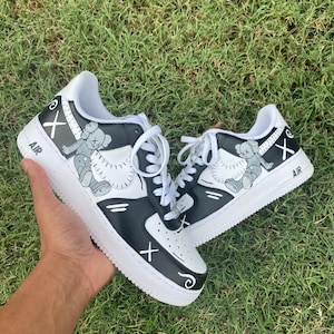 Peut inclure: Paire de baskets noires et blanches avec un motif de personnage de dessin anim&eacute;. Les chaussures ont des lacets blancs, une semelle blanche et le mot "AIR" est imprim&eacute; sur le c&ocirc;t&eacute;. Le design comprend un "X" et une figurine de dessin anim&eacute;.