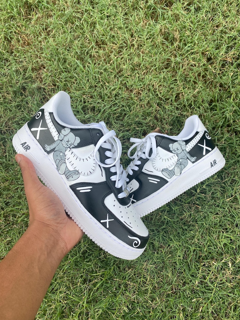 Peut inclure: Paire de baskets noires et blanches avec un motif de personnage de dessin anim&eacute;. Les chaussures ont des lacets blancs, une semelle blanche et le mot "AIR" est imprim&eacute; sur le c&ocirc;t&eacute;. Le design comprend un "X" et une figurine de dessin anim&eacute;.