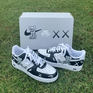 Peut inclure: Une paire de baskets Nike Air Force 1 blanches et noires avec des illustrations personnalis&eacute;es. Les baskets pr&eacute;sentent un dessin de personnage de dessin anim&eacute; et le logo Nike Air Force 1. Elles sont pos&eacute;es sur de l'herbe avec une bo&icirc;te blanche en arri&egrave;re-plan.