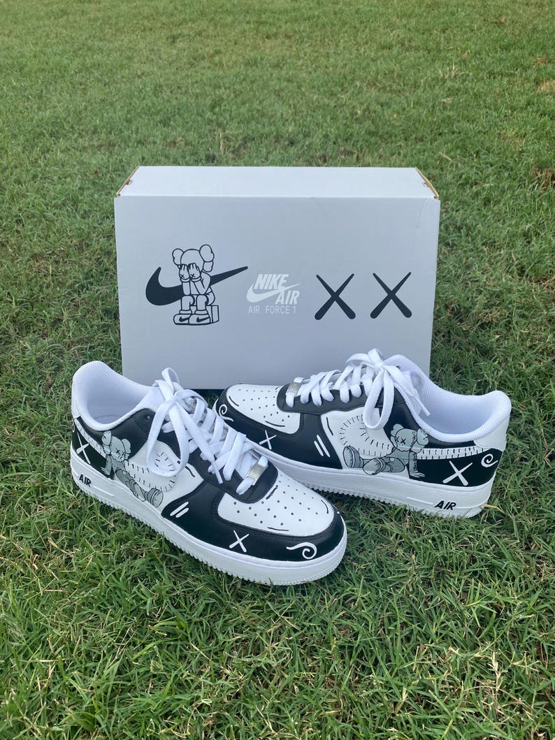 Peut inclure: Une paire de baskets Nike Air Force 1 blanches et noires avec des illustrations personnalis&eacute;es. Les baskets pr&eacute;sentent un dessin de personnage de dessin anim&eacute; et le logo Nike Air Force 1. Elles sont pos&eacute;es sur de l'herbe avec une bo&icirc;te blanche en arri&egrave;re-plan.