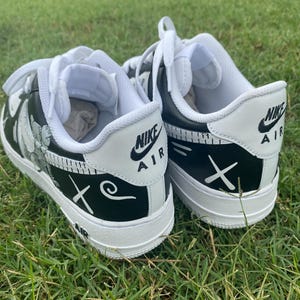 Peut inclure: Paire de baskets Nike Air Force 1 noires et blanches avec des motifs personnalis&eacute;s. Les chaussures ont des lacets, des semelles et des languettes de talon blancs avec le logo Nike Air. Les c&ocirc;t&eacute;s pr&eacute;sentent un motif floral et la lettre "X" et un tourbillon.