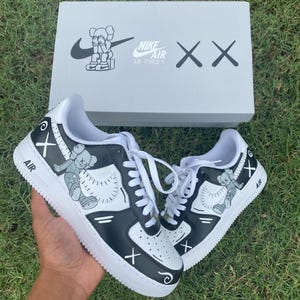 Peut inclure: Des baskets Nike Air Force 1 noires et blanches avec des illustrations personnalis&eacute;es et des lacets blancs. Le design comprend un personnage de dessin anim&eacute; et le logo Nike. Les chaussures sont pr&eacute;sent&eacute;es avec leur bo&icirc;te d'origine, qui porte &eacute;galement le logo Nike et les mots "Air Force 1".