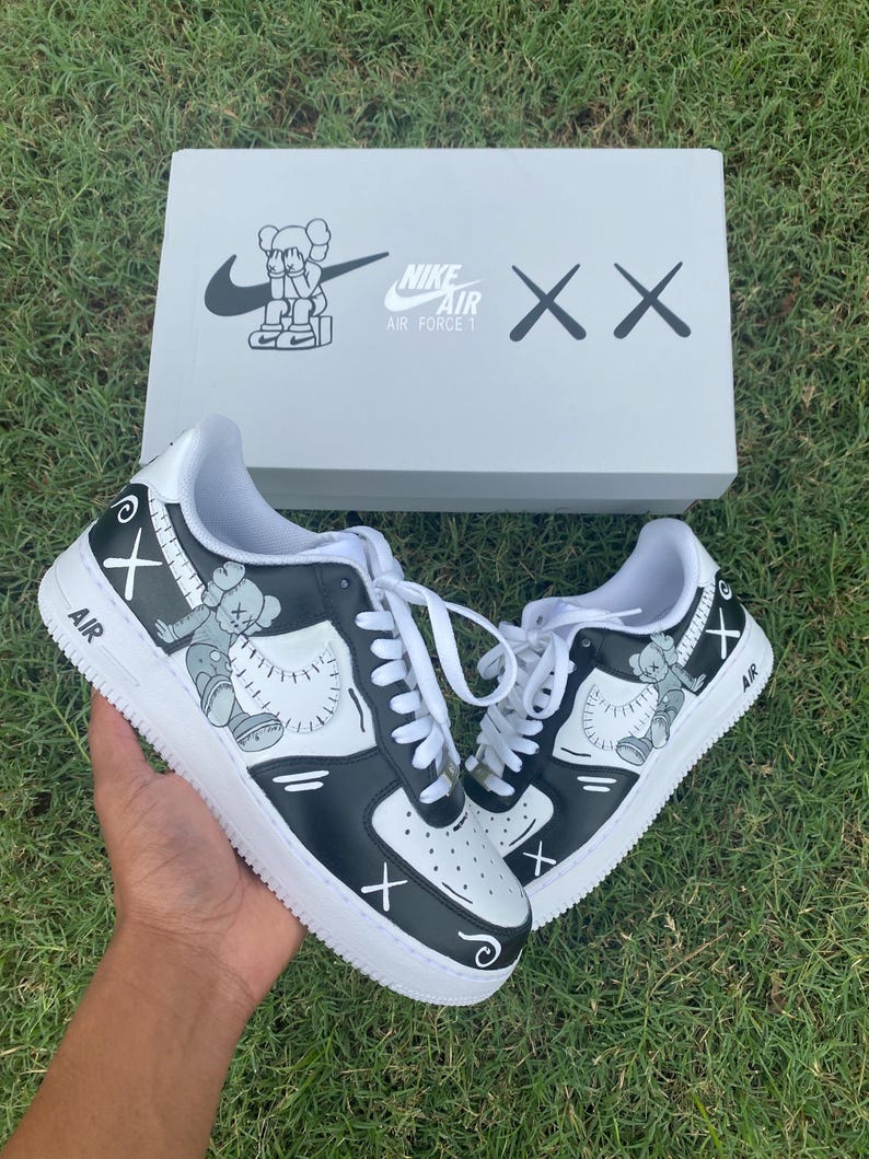 Peut inclure: Des baskets Nike Air Force 1 noires et blanches avec des illustrations personnalis&eacute;es et des lacets blancs. Le design comprend un personnage de dessin anim&eacute; et le logo Nike. Les chaussures sont pr&eacute;sent&eacute;es avec leur bo&icirc;te d'origine, qui porte &eacute;galement le logo Nike et les mots "Air Force 1".