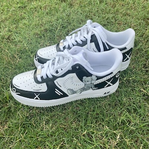Peut inclure: Paire de baskets blanches et noires avec des lacets blancs. Les baskets pr&eacute;sentent un dessin de personnage de dessin anim&eacute; sur le c&ocirc;t&eacute;, avec le mot "AIR" visible. Les chaussures sont pos&eacute;es sur de l'herbe verte.