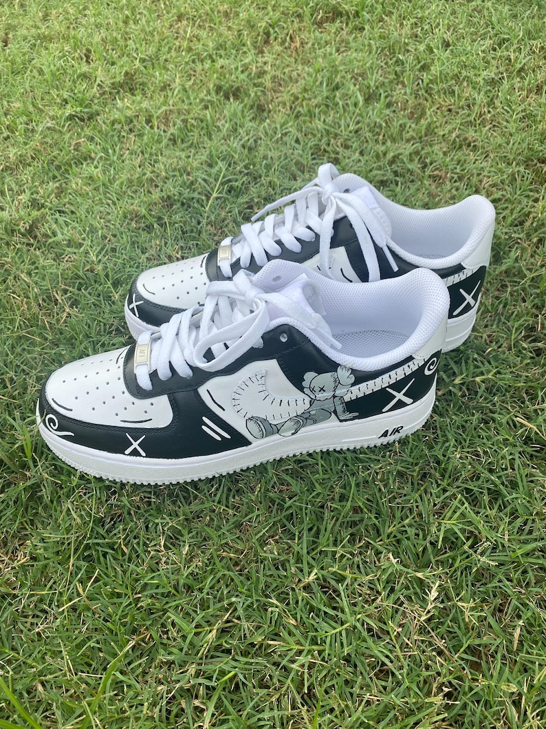Peut inclure: Paire de baskets blanches et noires avec des lacets blancs. Les baskets pr&eacute;sentent un dessin de personnage de dessin anim&eacute; sur le c&ocirc;t&eacute;, avec le mot "AIR" visible. Les chaussures sont pos&eacute;es sur de l'herbe verte.
