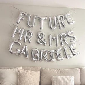 Könnte beinhalten: Silberne Folienballons in Buchstabenform, die in drei Reihen an einer Wand angeordnet sind und den Schriftzug FUTURE MR & MRS GABRIELE bilden. Die Ballons sind aufgeblasen und an dünnen Schnüren befestigt, als Dekoration für ein Fest.