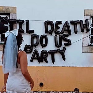 May include: A bride-to-be in a white dress and veil stands in front of a wall decorated with black balloons and a banner that reads "TIL DEATH DO US PARTY." The scene is set outdoors with cobblestone ground and a yellow and white wall.