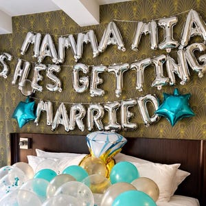 May include: Silver letter balloons spelling MAMMA MIA SHES GETTING MARRIED with two teal star balloons, a large diamond ring balloon, and a collection of clear, teal, and gold balloons on a bed with a pillow reading I found my HONEY.