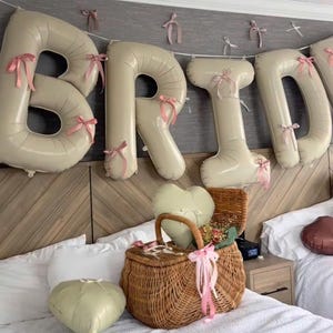 Peut inclure: Des ballons beiges en forme de lettres &eacute;pelant BRID, d&eacute;cor&eacute;s de petits n&oelig;uds en ruban rose, sont suspendus. Un panier en osier tress&eacute; marron est pos&eacute; sur un lit, contenant un ballon en forme de c&oelig;ur et des fleurs s&eacute;ch&eacute;es, avec un autre ballon c&oelig;ur.