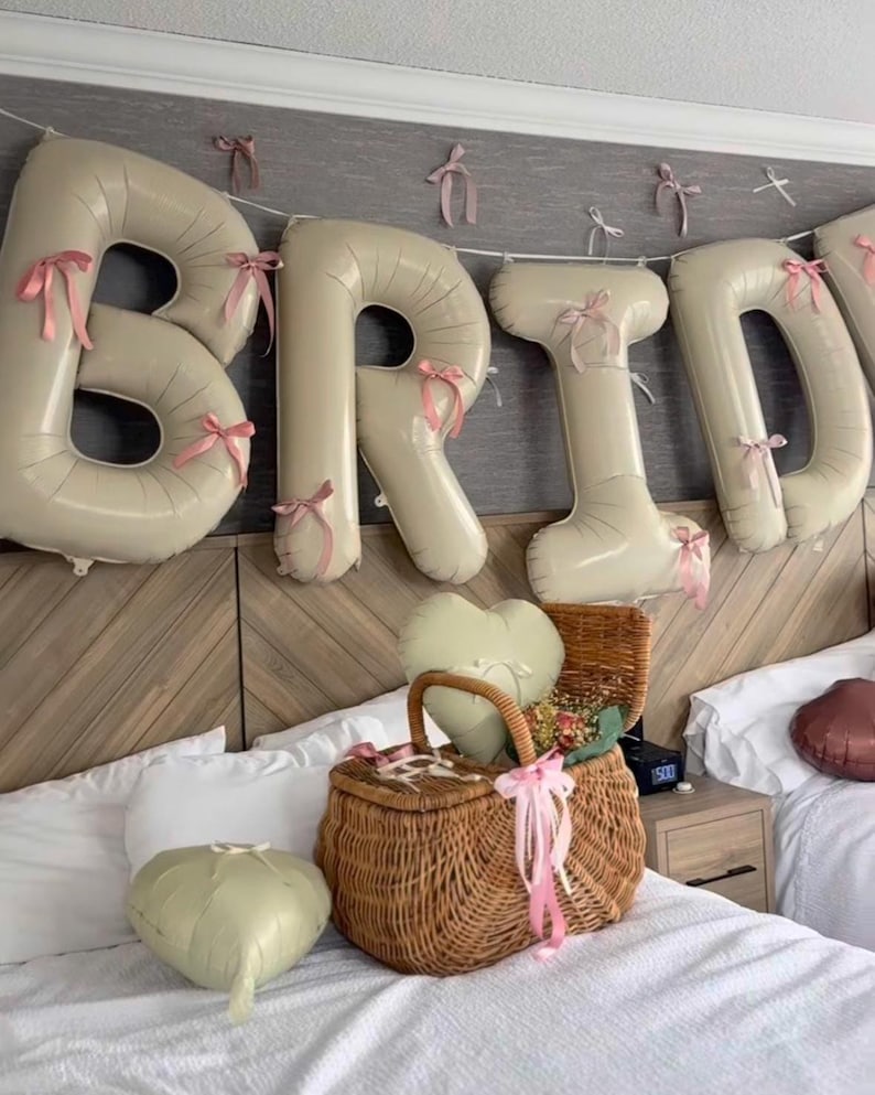 Peut inclure: Des ballons beiges en forme de lettres &eacute;pelant BRID, d&eacute;cor&eacute;s de petits n&oelig;uds en ruban rose, sont suspendus. Un panier en osier tress&eacute; marron est pos&eacute; sur un lit, contenant un ballon en forme de c&oelig;ur et des fleurs s&eacute;ch&eacute;es, avec un autre ballon c&oelig;ur.